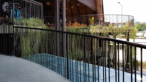 A modern outdoor space with a curved balcony railing overlooking a lower level. Hanging plants cascade over the edge, adding greenery to the industrial design. In the background, there are wooden chairs and tables under a high ceiling structure. Some bicycles can be seen parked to the side.