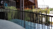 A modern outdoor space with a curved balcony railing overlooking a lower level. Hanging plants cascade over the edge, adding greenery to the industrial design. In the background, there are wooden chairs and tables under a high ceiling structure. Some bicycles can be seen parked to the side.