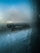 Condensation covers a glass window, with droplets of water creating a misty, blurred effect. The background is barely visible through the moisture, displaying a hint of a landscape with pale blue sky and horizon.