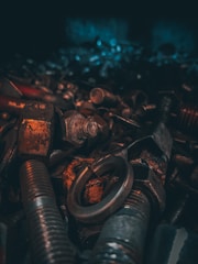 A collection of various metal bolts, nuts, and washers is shown in a dimly lit environment. The objects exhibit signs of rust and aging, with a focus on texture and detail.