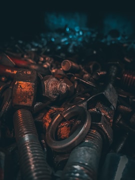 A collection of various metal bolts, nuts, and washers is shown in a dimly lit environment. The objects exhibit signs of rust and aging, with a focus on texture and detail.