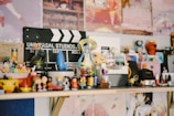 A collection of various figurines and small toys is arranged on a shelf. There's a Universal Studios clapperboard in the background along with colorful posters. The figurines include diverse characters from different media, arranged in a lively and eclectic manner.