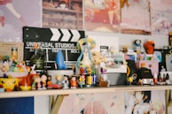 A collection of various figurines and small toys is arranged on a shelf. There's a Universal Studios clapperboard in the background along with colorful posters. The figurines include diverse characters from different media, arranged in a lively and eclectic manner.