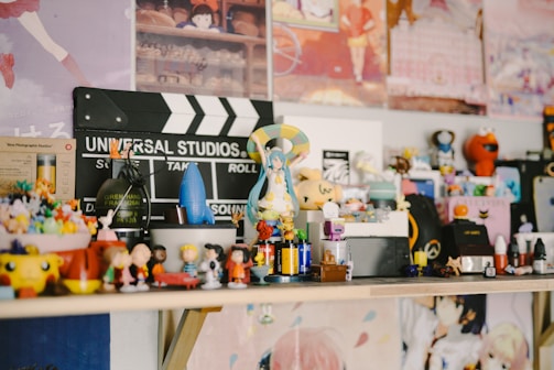A collection of various figurines and small toys is arranged on a shelf. There's a Universal Studios clapperboard in the background along with colorful posters. The figurines include diverse characters from different media, arranged in a lively and eclectic manner.