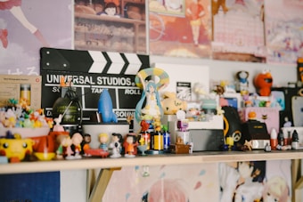 A collection of various figurines and small toys is arranged on a shelf. There's a Universal Studios clapperboard in the background along with colorful posters. The figurines include diverse characters from different media, arranged in a lively and eclectic manner.