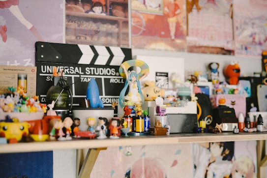 A collection of various figurines and small toys is arranged on a shelf. There's a Universal Studios clapperboard in the background along with colorful posters. The figurines include diverse characters from different media, arranged in a lively and eclectic manner.