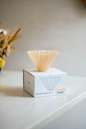 A light-colored, fluted coffee dripper sits on top of a white box labeled 'Origami Dripper Air S'. The box rests on a light wooden surface, with blurred flowers in the background.