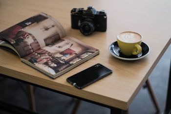 A wooden table holds an open magazine featuring interior photographs, a vintage black camera, a smartphone, and a cup of latte with art in a yellow cup on a saucer.