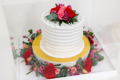 A beautifully decorated white cake with a textured icing pattern is topped with a floral arrangement of red and pink flowers along with green leaves. The cake sits on a gold base and is surrounded by a wreath of similar flowers and leaves.