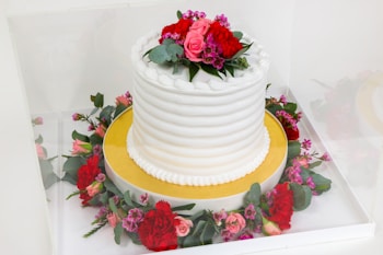 A beautifully decorated white cake with a textured icing pattern is topped with a floral arrangement of red and pink flowers along with green leaves. The cake sits on a gold base and is surrounded by a wreath of similar flowers and leaves.