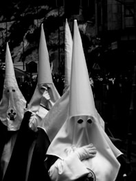 A group of individuals dressed in white robes with tall pointed hoods, covering their faces, are walking in a procession. The setting appears to be a street with buildings in the background, and the image is in black and white.