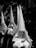 A group of individuals dressed in white robes with tall pointed hoods, covering their faces, are walking in a procession. The setting appears to be a street with buildings in the background, and the image is in black and white.