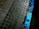 Weathered, square tiles set in a grid pattern make up a paved surface with a partially painted curb. The curb is painted in blue, but the paint is chipped and worn, exposing the aged, rusted surface underneath.