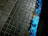 Weathered, square tiles set in a grid pattern make up a paved surface with a partially painted curb. The curb is painted in blue, but the paint is chipped and worn, exposing the aged, rusted surface underneath.