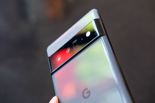 A close-up of a modern smartphone featuring a sleek design with a glossy finish, showcasing a distinctive horizontal camera bar near the top. The phone's reflective back partially displays the Google logo, set against a blurred background.