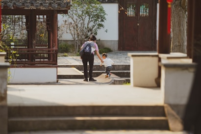 A person with a backpack helps a young child step up onto a raised stone platform in a traditional garden setting. The environment features a wooden structure with intricate lattice work, surrounded by greenery and a stone pathway leading to a wooden door.