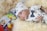 A baby wearing a white outfit adorned with small blue patterns and a matching hat is lying on a fluffy cream-colored blanket. The outfit features a blue polka-dot bowtie and decorative pins. In the background, there are colorful toys and a blurred packaging box.