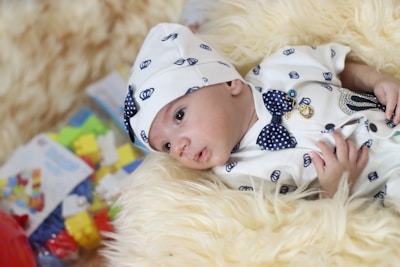 A baby wearing a white outfit adorned with small blue patterns and a matching hat is lying on a fluffy cream-colored blanket. The outfit features a blue polka-dot bowtie and decorative pins. In the background, there are colorful toys and a blurred packaging box.