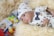 A baby wearing a white outfit adorned with small blue patterns and a matching hat is lying on a fluffy cream-colored blanket. The outfit features a blue polka-dot bowtie and decorative pins. In the background, there are colorful toys and a blurred packaging box.