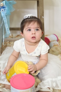 A young child wearing a white dress sits on the floor holding a yellow canister. Behind the child, there are several other canisters with pink and red lids. To the left, a blue ribbon decorates a piece of furniture.