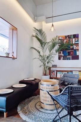 A cozy corner in a cafe with a bohemian vibe. A large tropical plant in a pot adds greenery to the room. A wooden barrel labeled 'Blue Mountain Coffee Product of Jamaica W.I.' serves as a table. There are patterned chairs and a black leather bench with circular white trays. The floor is decorated with a round tribal-patterned rug. A stylish pendant light hangs from the ceiling, and there's a framed whimsical artwork on the wall.