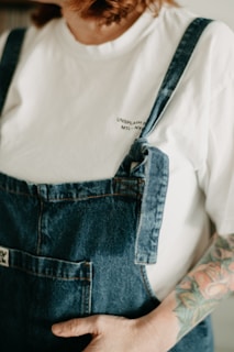 A person wearing a white t-shirt with a text print partially visible under blue denim overalls. They have a visible arm tattoo featuring colorful floral designs and their hand is placed in the pocket of the overalls. The overall image has a soft, warm appearance from the lighting.