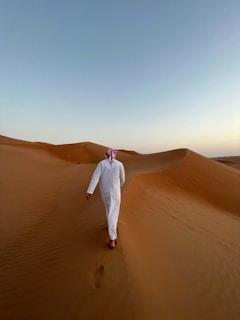 A person wearing a traditional white thobe and a headscarf walks through a vast desert landscape characterized by smooth sand dunes and a clear sky above.