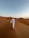 A person wearing a traditional white thobe and a headscarf walks through a vast desert landscape characterized by smooth sand dunes and a clear sky above.