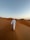 A person wearing a traditional white thobe and a headscarf walks through a vast desert landscape characterized by smooth sand dunes and a clear sky above.
