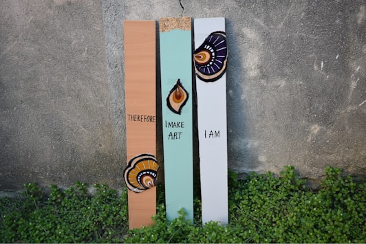 Three vertical wooden planks stand against a textured concrete wall. Each plank is painted with a unique decoration and text. The left plank is brown with the word 'THEREFORE' and an artistic design at the bottom. The middle plank is light green with a geometric pattern and the words 'I MAKE ART'. The right plank is pale gray with the words 'I AM' and another abstract design near the top. Small green plants grow at the base of the planks.