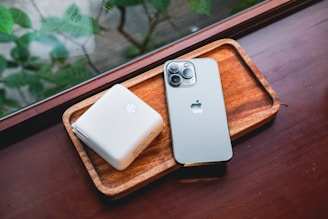 A wooden tray holds a smartphone with a metallic finish and a white square device, possibly a charger or power bank. The items rest on a polished wooden surface, and the background shows a blurred view of greenery through a window.