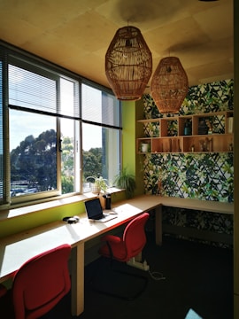 A small home office with two red chairs and a long wooden desk placed next to large windows that allow natural light to illuminate the space. There are two woven pendant lights hanging from the ceiling, and a laptop sits open on the desk. A vibrant green plant is positioned near the window, and a patterned accent wall features a mix of green and blue tones with a bookshelf mounted on it.