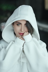 A person wearing a white hooded sweatshirt is gazing directly at the camera, with a calm and introspective expression. The hood partially covers their head, and they have dark hair peeking through. Their hands are up near their face, creating a cozy and relaxed look, with a blurred background that enhances the focus on the central figure.