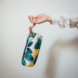 A hand is holding a colorful pouch with a leaf pattern design. The pouch has a light brown strap, and the background is a plain, neutral color.