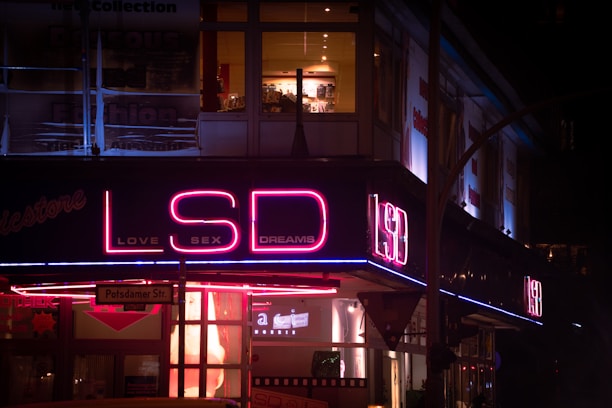 A neon sign displaying the words 'LSD' in large letters with 'LOVE SEX DREAMS' underneath. The sign is illuminated in pink and blue neon lights, attached to a building on Potsdamer Str. The surrounding area appears to be a nighttime city scene with visible storefronts and windows.