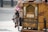 A traditional street barrel organ with ornate woodwork and painted illustrations. A toy monkey, adorned with a pink feathered hat and a small vest, is attached to the organ, holding a metal container.