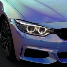 A close-up view of a car's front section showcasing sleek, modern design. The headlight is prominent, with intricate LED detailing that appears illuminated. The surface reflects a subtle gradient of colors, including blue and purple hues. The alloy wheel is stylishly designed with multiple spokes.