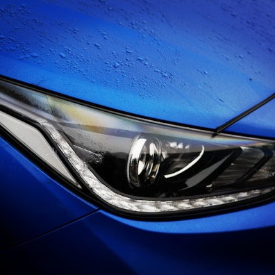 A close-up view of a car's headlight, showcasing intricate design elements and sleek curves. The surface has droplets of water, suggesting recent rain or a wash. The headlight is modern and features sharp, angular lines with a reflective surface.