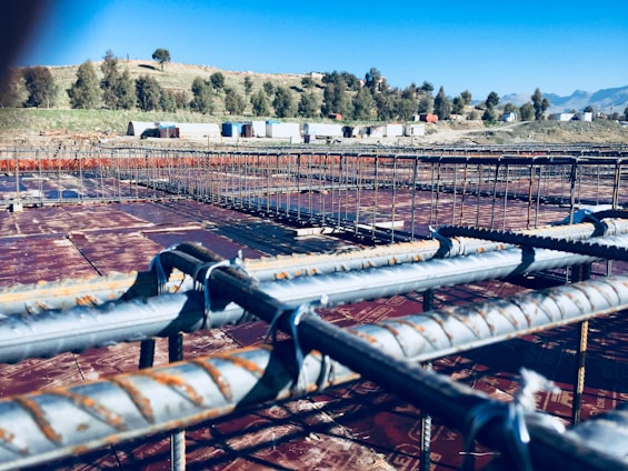 A construction site features a grid of rusted steel rebar arranged in preparation for concrete pouring. Beyond the framework, trees and small portable buildings are visible against a clear blue sky, with hills in the distant background.