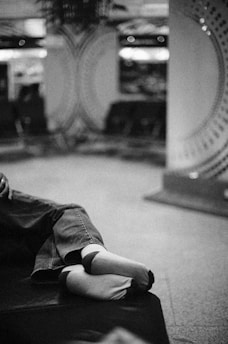 A pair of feet wearing socks and jeans rest on a cushioned surface in a blurred indoor setting. The background features circular patterns with soft lighting, suggesting a relaxed environment.