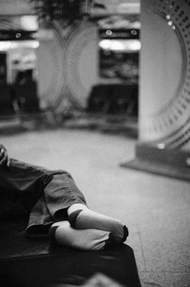 A pair of feet wearing socks and jeans rest on a cushioned surface in a blurred indoor setting. The background features circular patterns with soft lighting, suggesting a relaxed environment.