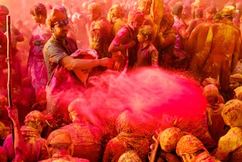 A large crowd of people is gathered, all covered in vibrant colors, specifically red and pink. They are taking part in a lively and energetic festival where colored powders are being thrown and spread around. The atmosphere is busy and festive, with people smiling and interacting closely.
