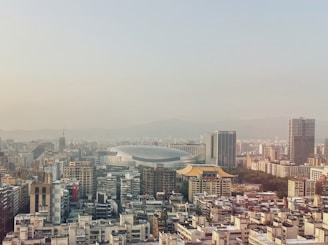 A cityscape featuring a mix of modern and traditional architecture. Skyscrapers dominate the landscape, with a large, dome-shaped sports stadium in the center. Traditional-style roofs with ornate designs are visible among the buildings. The background reveals a mountain range under a hazy sky.
