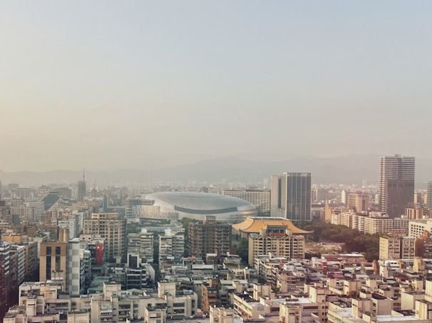 A cityscape featuring a mix of modern and traditional architecture. Skyscrapers dominate the landscape, with a large, dome-shaped sports stadium in the center. Traditional-style roofs with ornate designs are visible among the buildings. The background reveals a mountain range under a hazy sky.