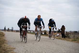 Three cyclists are riding vintage road bicycles on a rural path, with a woman in a brown coat taking a photo or observing them. The cyclists appear to be participating in a race or an event, dressed in cycling gear suitable for cooler weather. The background shows an expansive landscape with some houses and trees under an overcast sky.