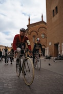 Two cyclists ride vintage bicycles on a cobblestone street in front of a historic brick building with Gothic architecture. The lead cyclist wears a red sweater, goggles, and a numbered armband, suggesting participation in a cycling event. Several pigeons are on the street while people walk in the background, creating a lively urban scene.