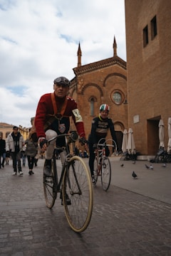 Two cyclists ride vintage bicycles on a cobblestone street in front of a historic brick building with Gothic architecture. The lead cyclist wears a red sweater, goggles, and a numbered armband, suggesting participation in a cycling event. Several pigeons are on the street while people walk in the background, creating a lively urban scene.