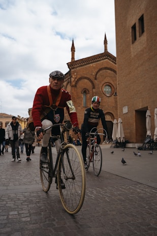Two cyclists ride vintage bicycles on a cobblestone street in front of a historic brick building with Gothic architecture. The lead cyclist wears a red sweater, goggles, and a numbered armband, suggesting participation in a cycling event. Several pigeons are on the street while people walk in the background, creating a lively urban scene.