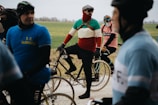 Cyclists are gathered on a road with vintage-style bicycles, wearing colorful outfits that suggest a retro theme. The participants appear to be in a relaxed setting with an overcast sky, indicating a cycling event in a rural area with green fields in the background.