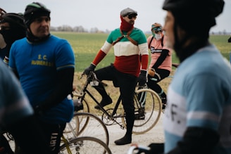 Cyclists are gathered on a road with vintage-style bicycles, wearing colorful outfits that suggest a retro theme. The participants appear to be in a relaxed setting with an overcast sky, indicating a cycling event in a rural area with green fields in the background.